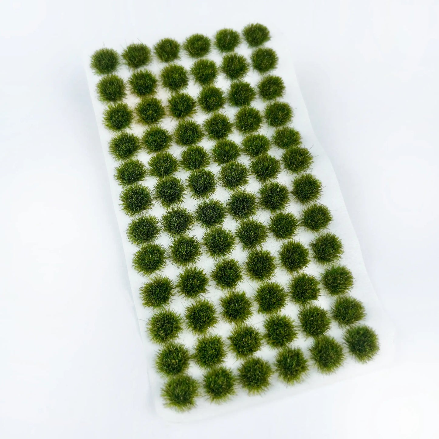 Unpacked sheet of dark forest green self-adhesive static grass tufts on clean white background, dense fine fiber bush clumps for military diorama, wargame terrain and scale railway scene landscaping