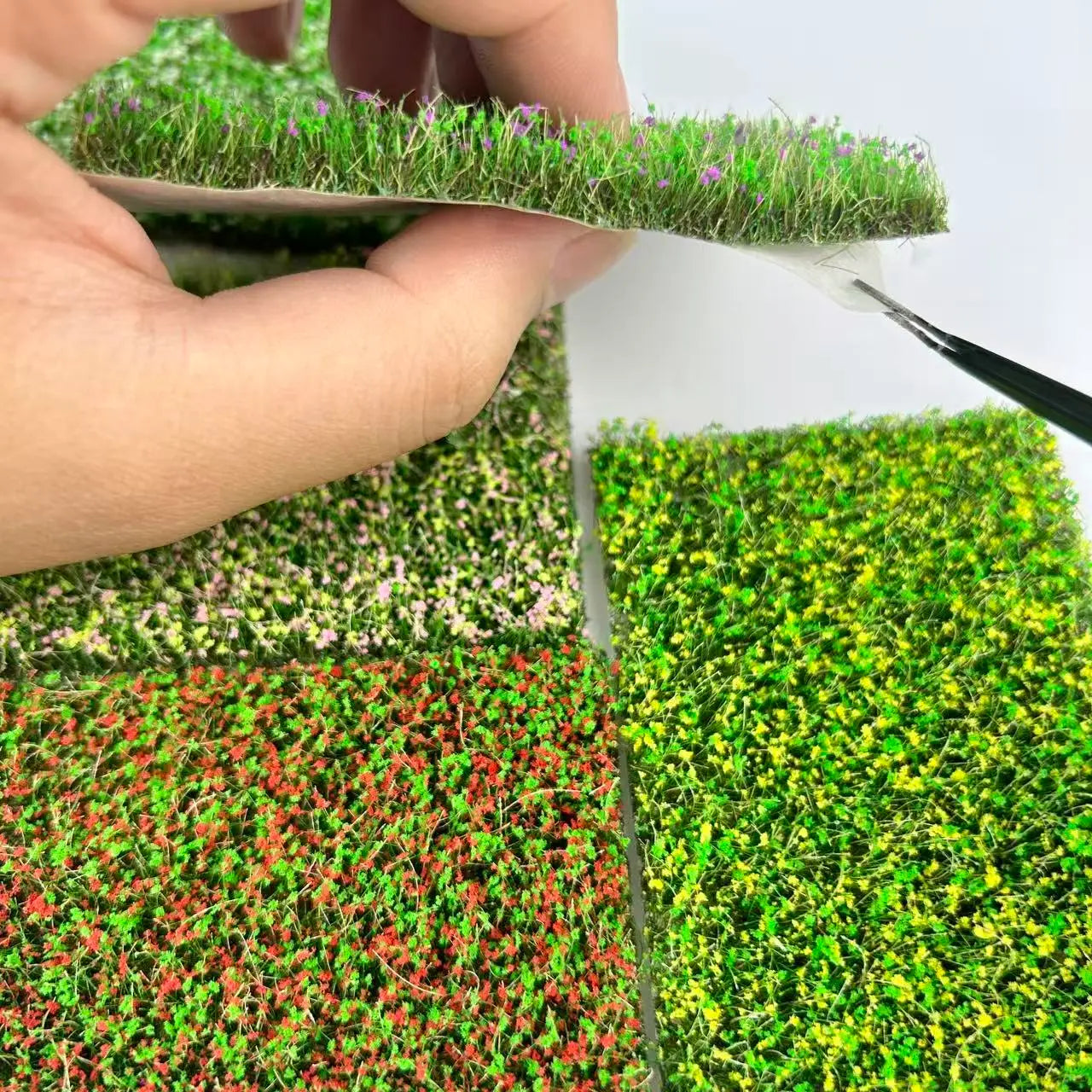 Hand lifting self adhesive artificial grass mat with purple flowers, showing four color options including purple, pink, red and yellow for diorama miniature terrain craft