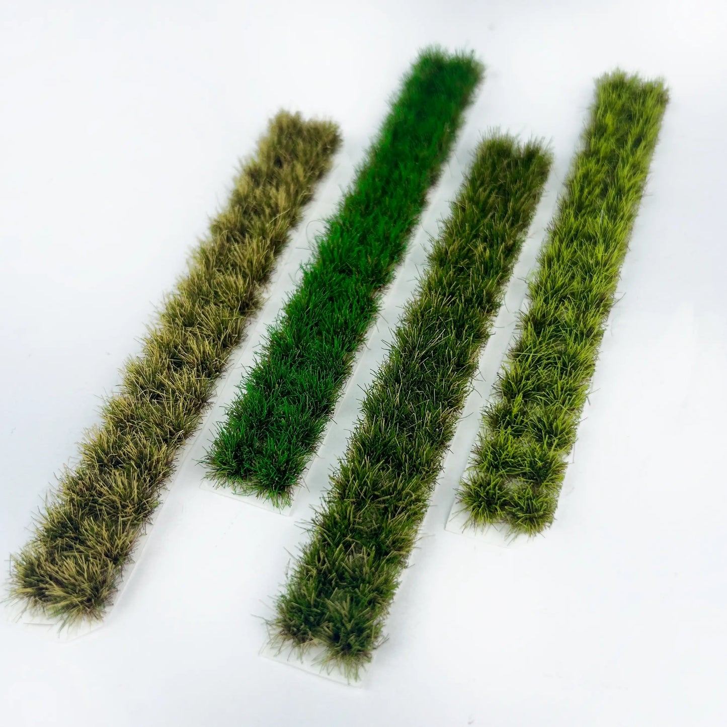 Four individual long miniature static grass tuft strips in mixed green and dry brown tones, marked with full size measurements for diorama and wargame terrain craft reference
