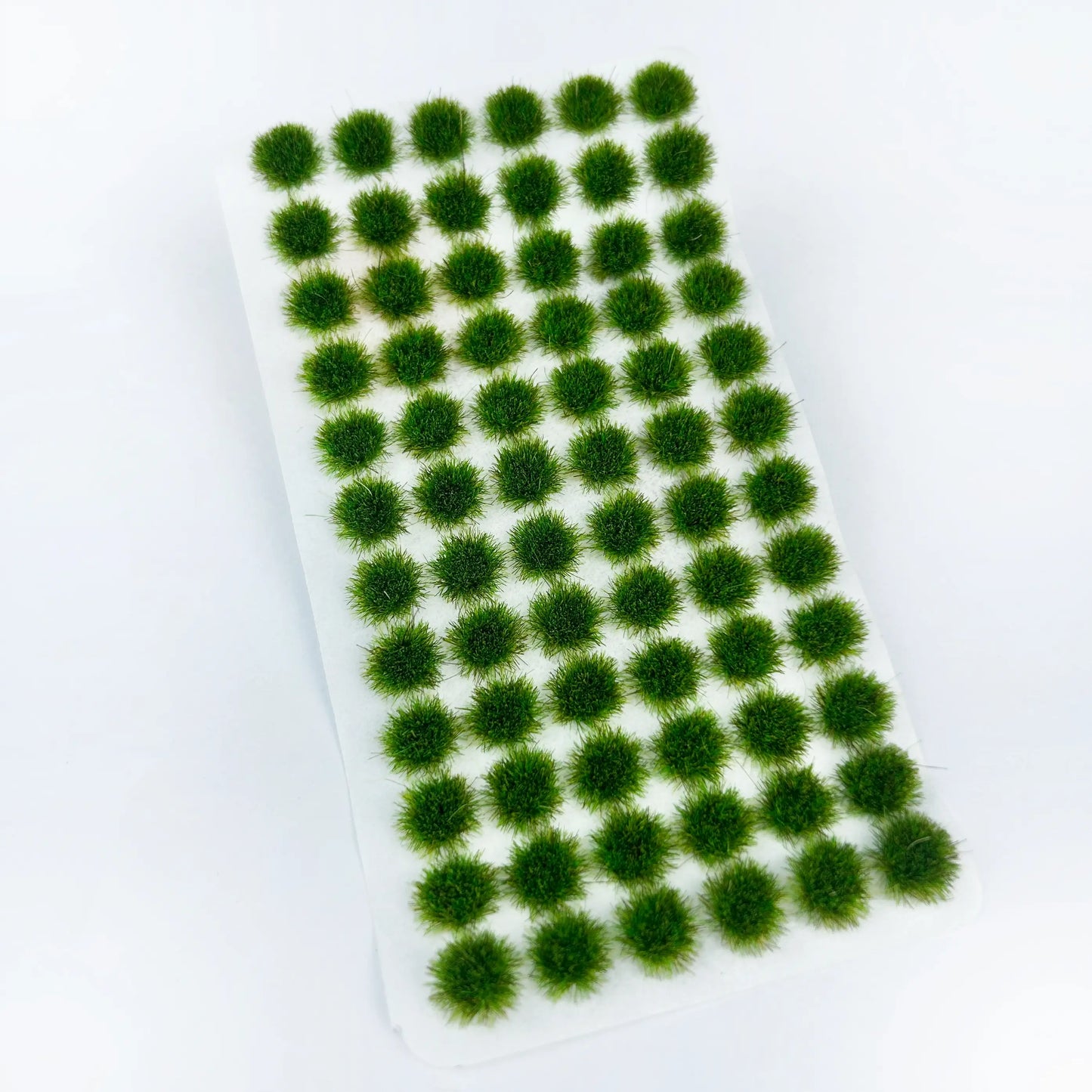 Unpacked sheet of dark forest green self-adhesive static grass tufts on clean white background, dense fine fiber bush clumps for military diorama, wargame terrain and scale railway scene landscaping