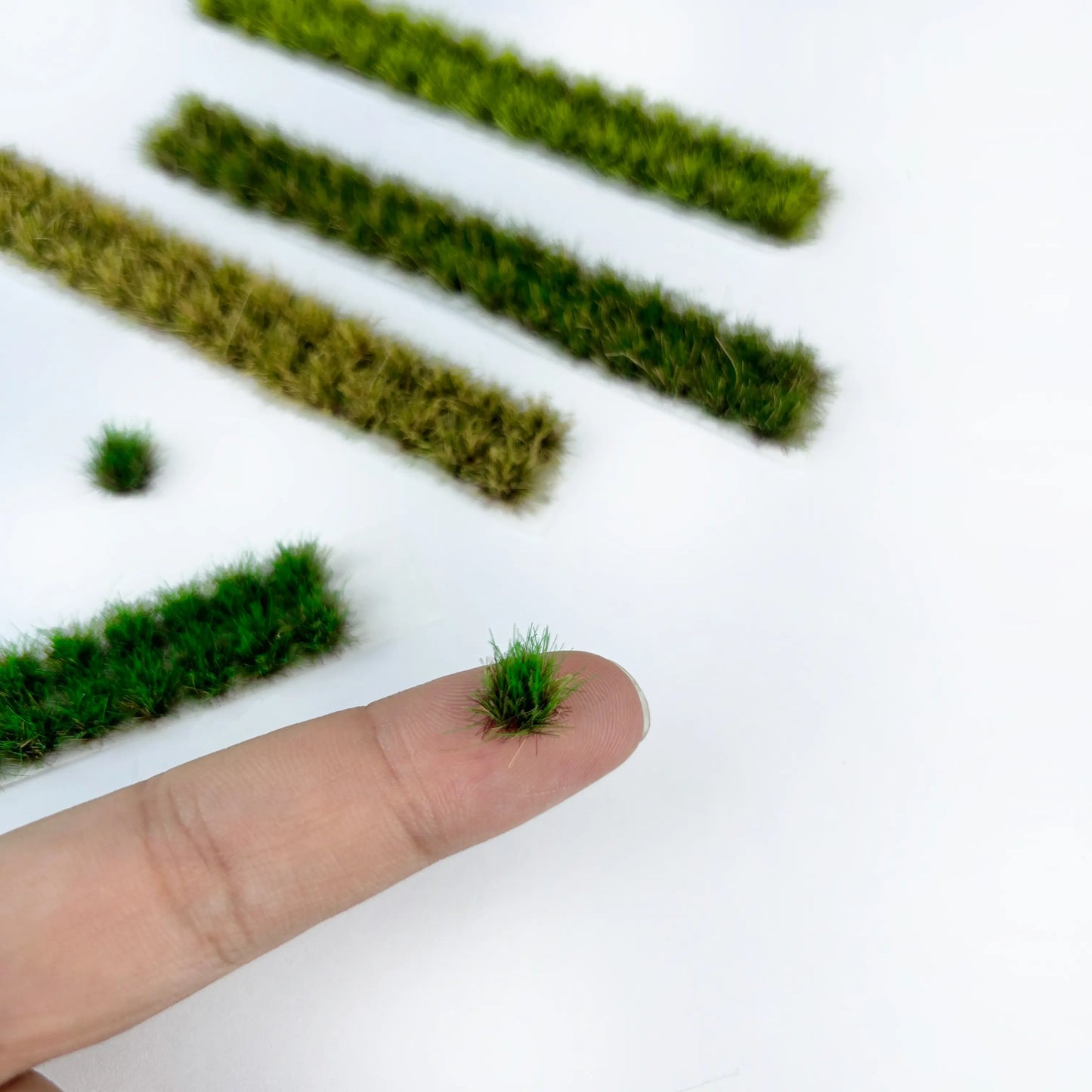 Tiny self-adhesive static grass tufts and long grass strips displayed on fingertip, marked with size measurements for miniature diorama terrain craft reference