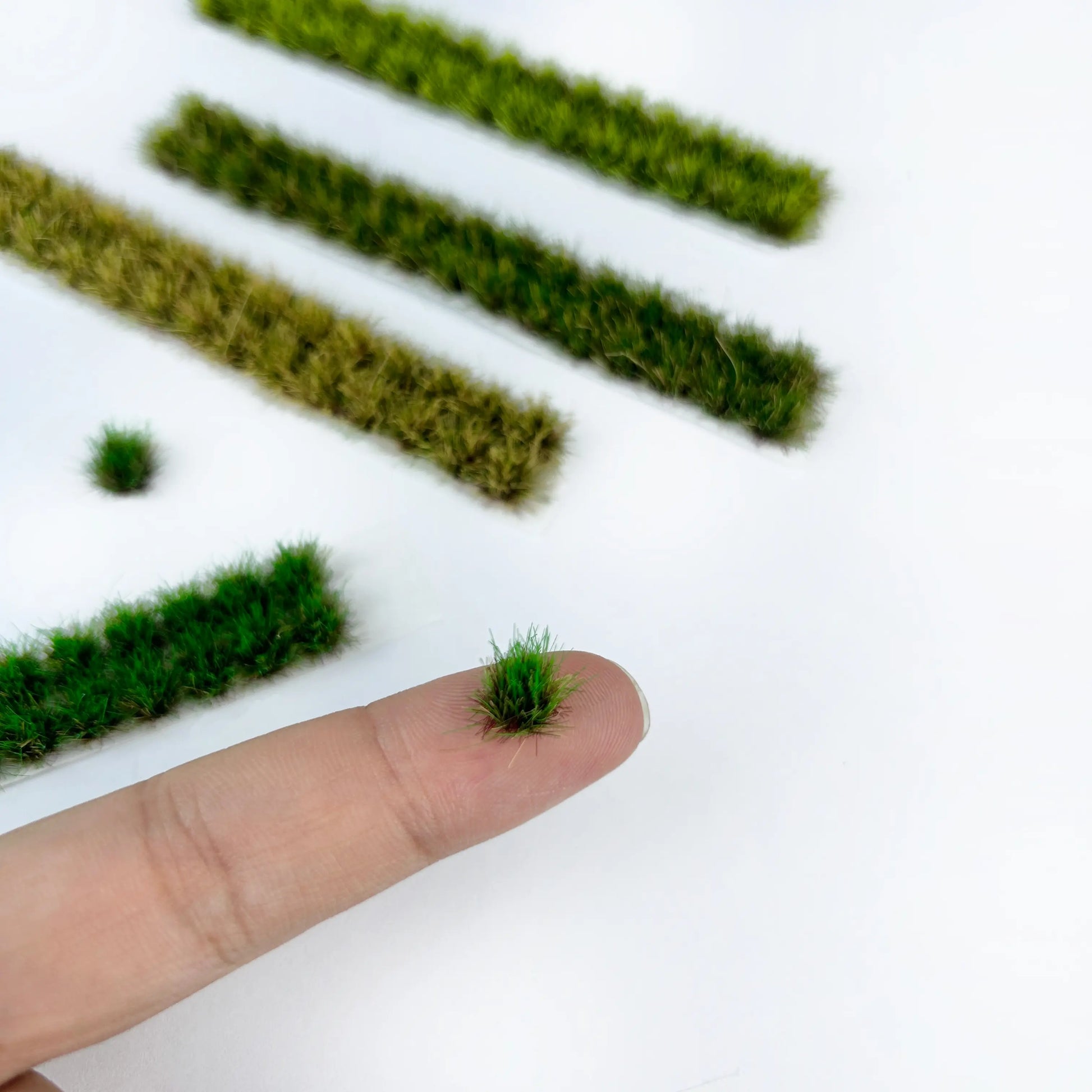 Tiny self-adhesive static grass tufts and long grass strips displayed on fingertip, marked with size measurements for miniature diorama terrain craft reference