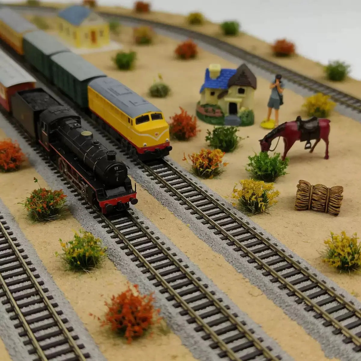 Scale model railroad layout diorama featuring steam & diesel locomotives, tiny cottage, mini human figure, red horse, assorted static grass tuft foliage and hay bale terrain decorations for wargame & train scenery making