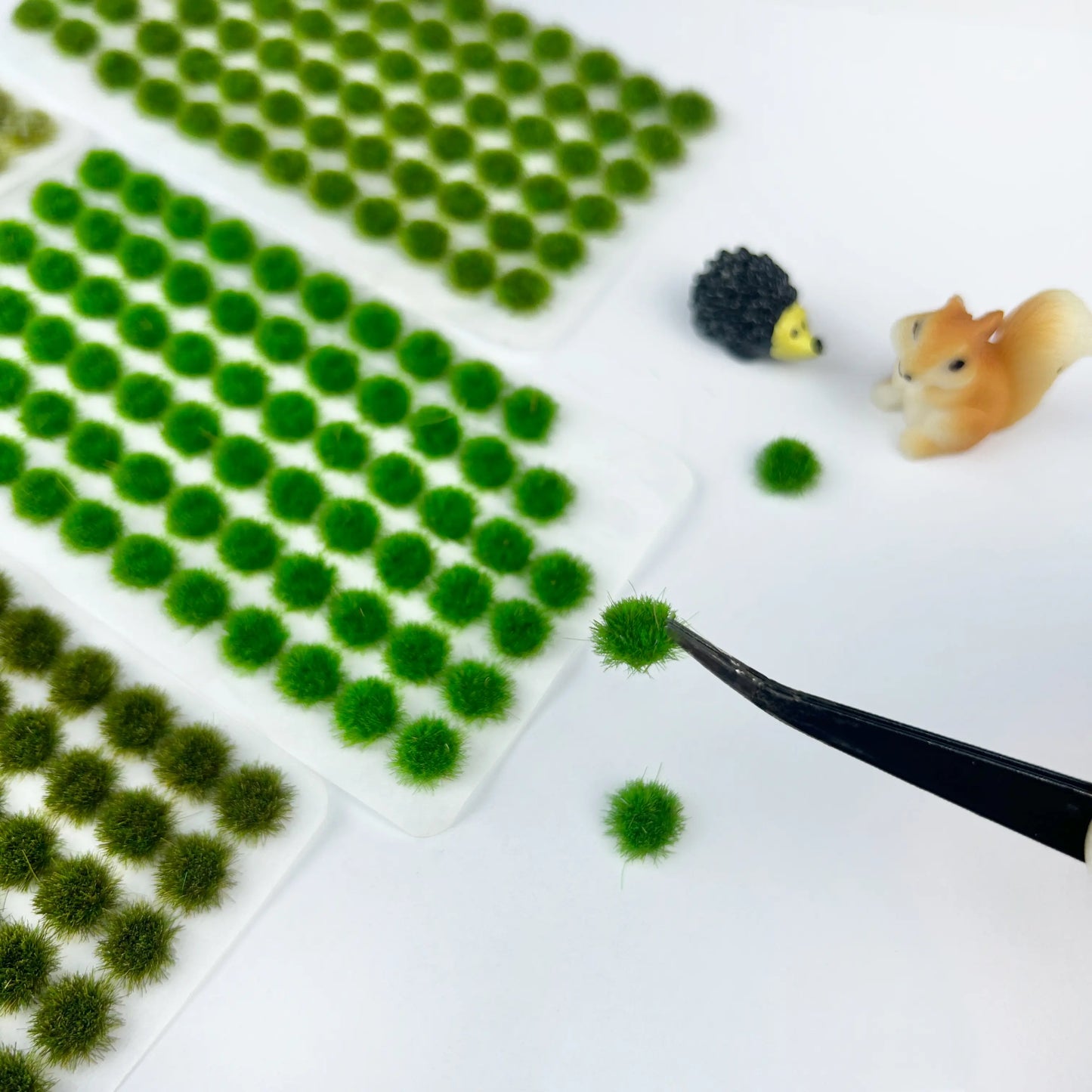 Craft demonstration scene with assorted color static grass tuft sheets, tweezers picking single grass clump, hedgehog and squirrel mini figures, countryside miniature diorama making reference display