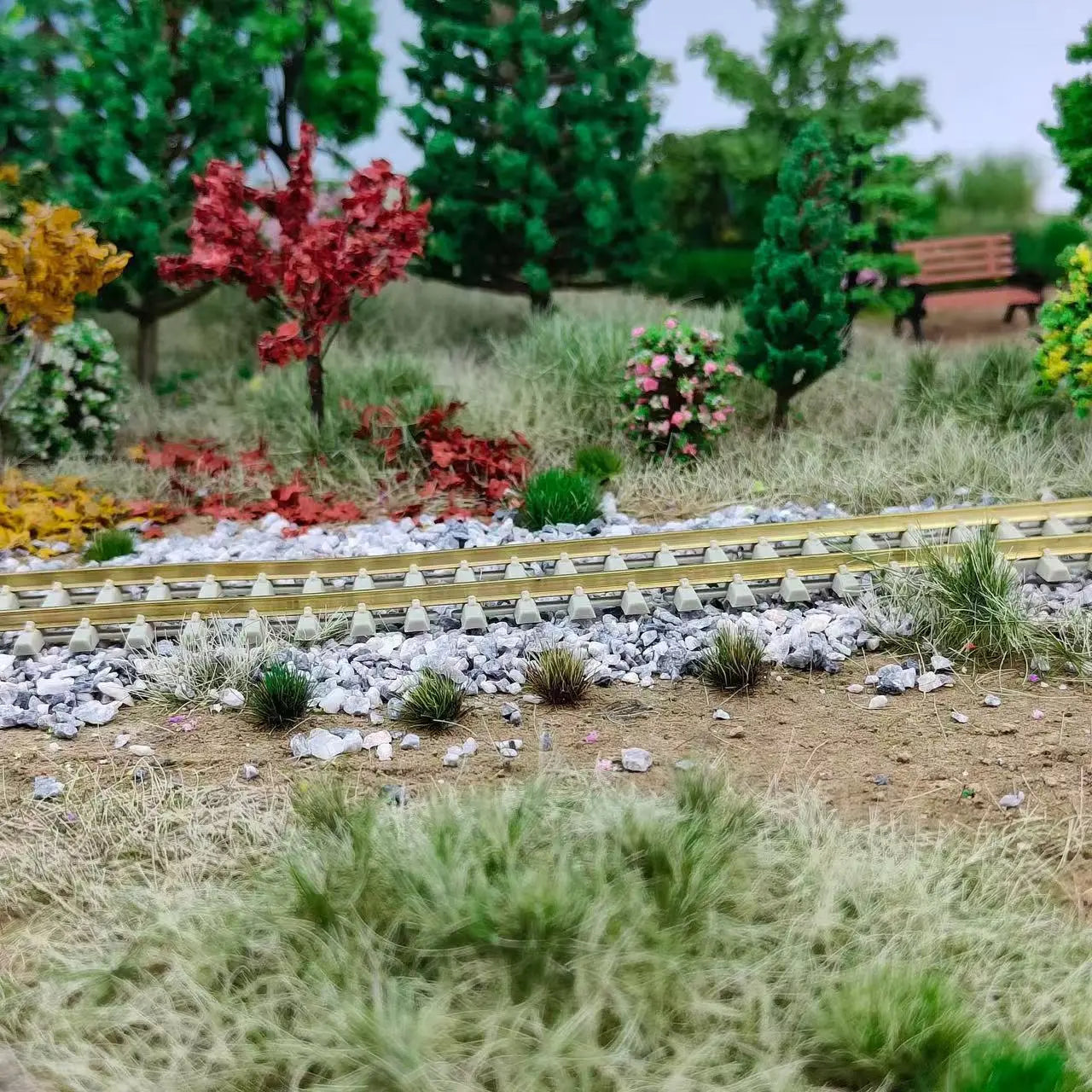 Miniature railway layout diorama with assorted static grass tufts, colorful model trees and gravel tracks, marked with full size measurements for train terrain craft reference