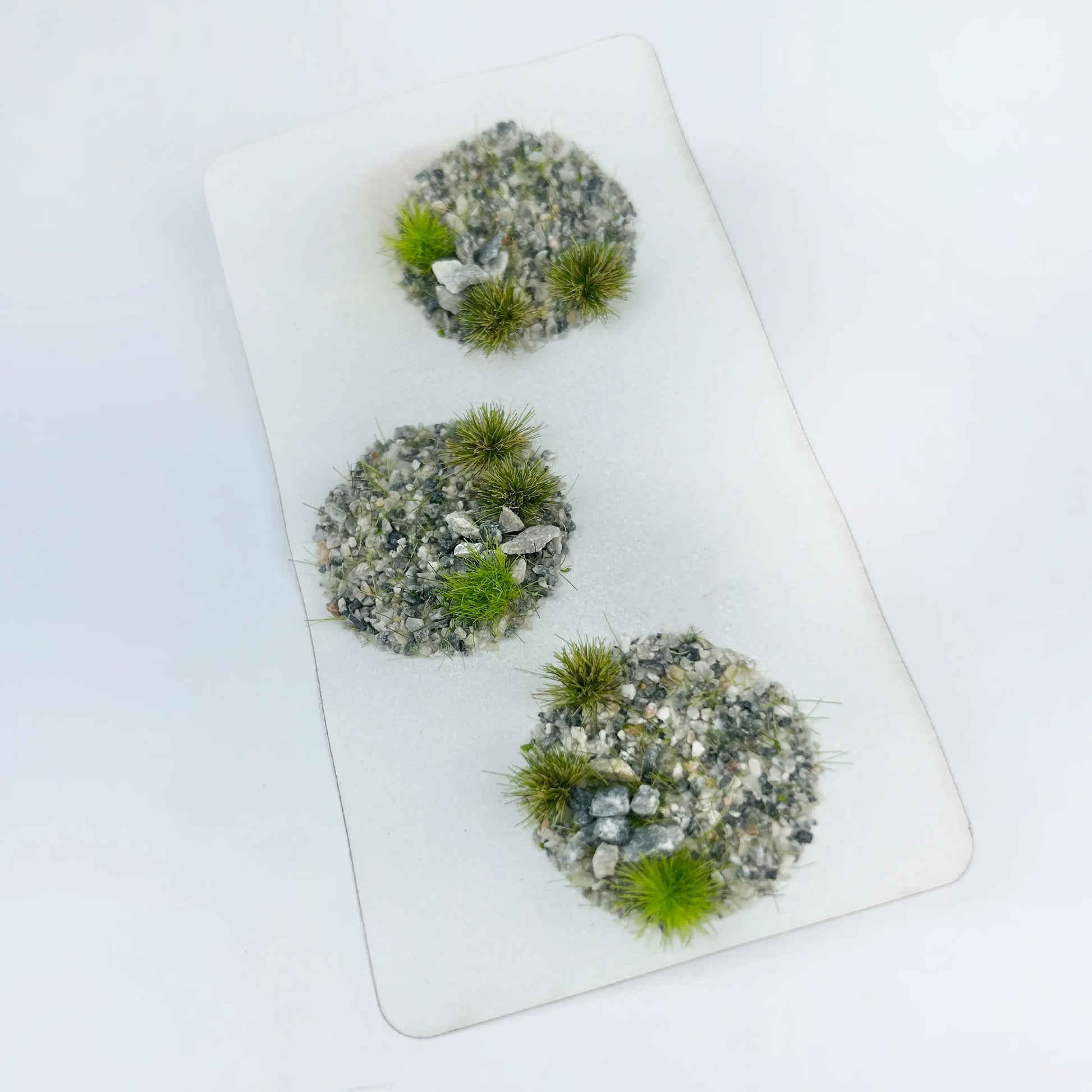 Unpainted wargame mini bases decorated with self-adhesive gravel pebble grass tufts, rocky terrain foliage samples for custom warhammer, historical and fantasy miniature model basing

